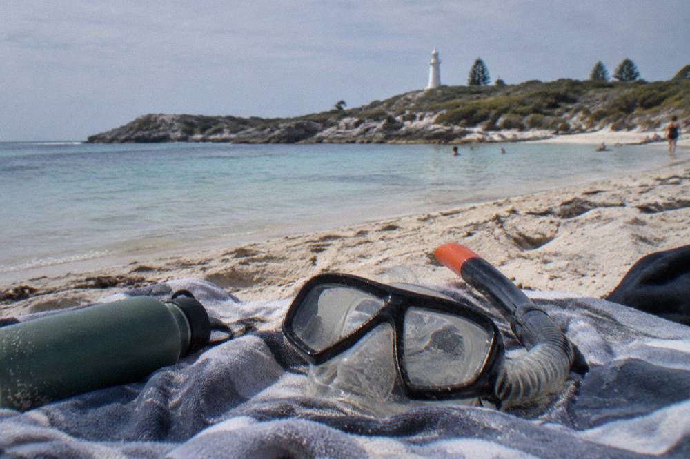 A snorkel and mask resting on a towel beside calm turquoise water at a Rottnest bay