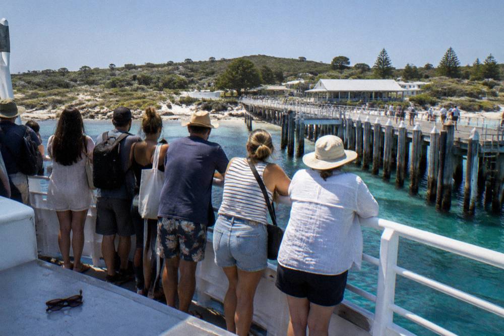 Crowded ferry deck with passengers in sunglasses looking out over open water toward an island