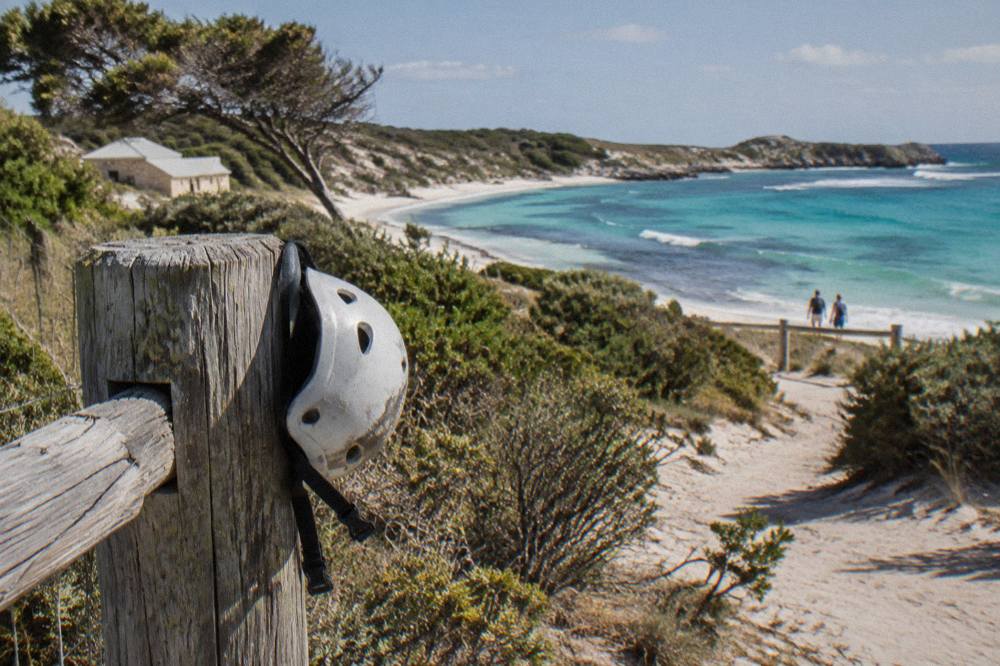 Bicycles parked on a sandy path beside a low coastal fence and clear water