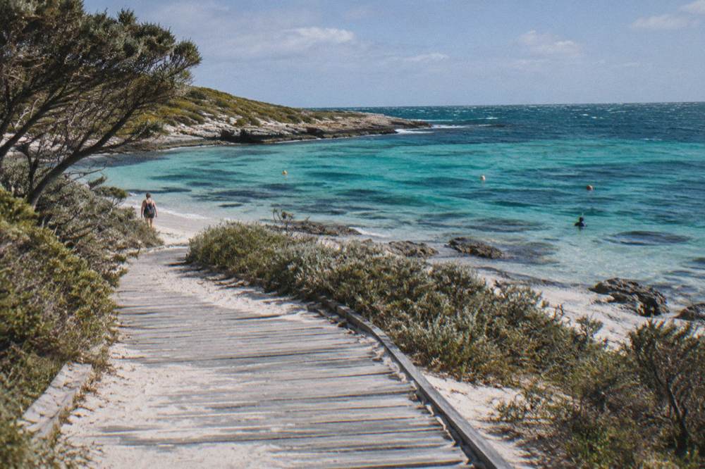 A wooden boardwalk over dunes leading toward a sheltered bay with snorkel buoys visible