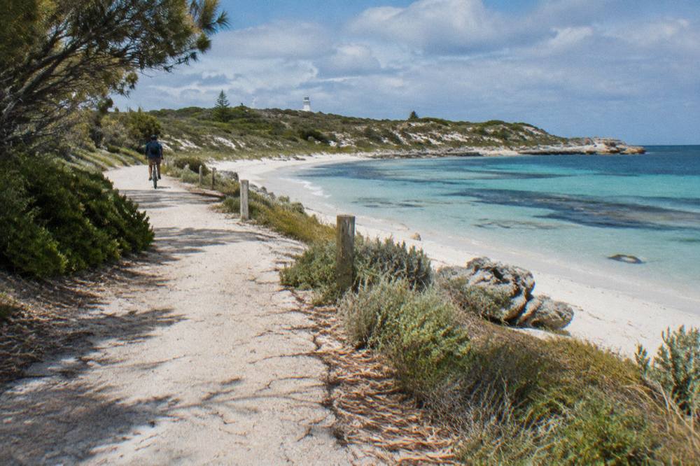 A cyclist riding a path beside white sand and clear shallow water at a Rottnest bay