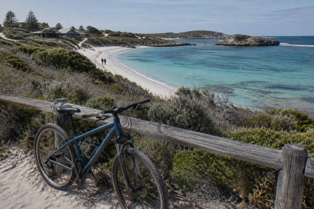 A bicycle leaning against a wooden fence beside a sandy path near turquoise island water