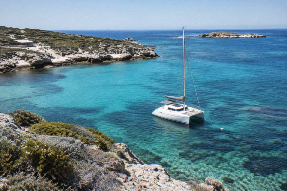 A small sailing catamaran moored over clear shallow reef water near a rocky headland