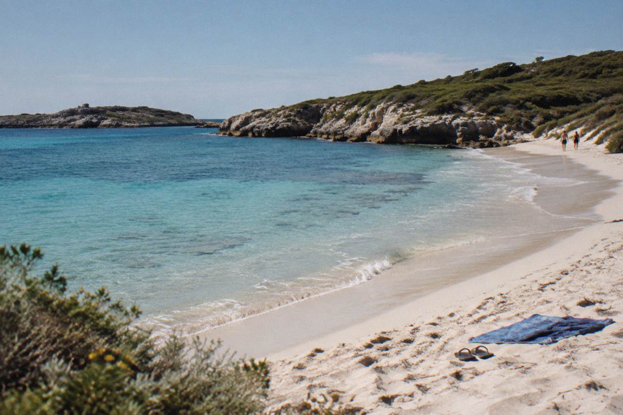 A calm bay on Rottnest Island with turquoise water lapping a pale sand beach and low coastal scrub