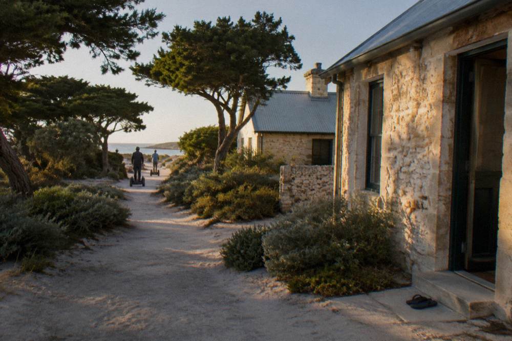 A quiet sandy path lined with low coastal scrub leading away from the settlement buildings
