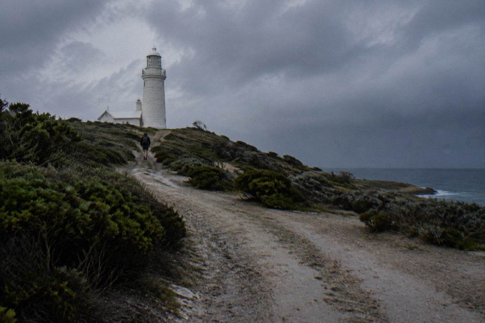 A rider walking a bicycle up a gravel path toward a white lighthouse above low coastal scrub