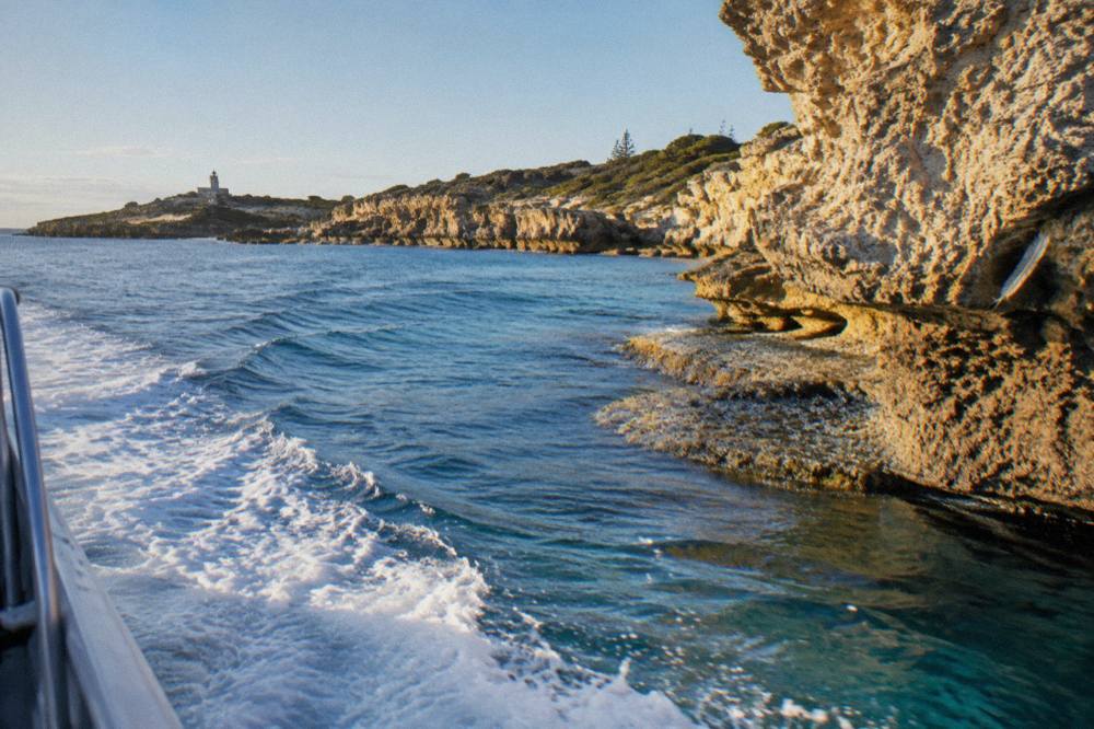 A cruising boat cutting across calm blue water near a rocky Rottnest headland
