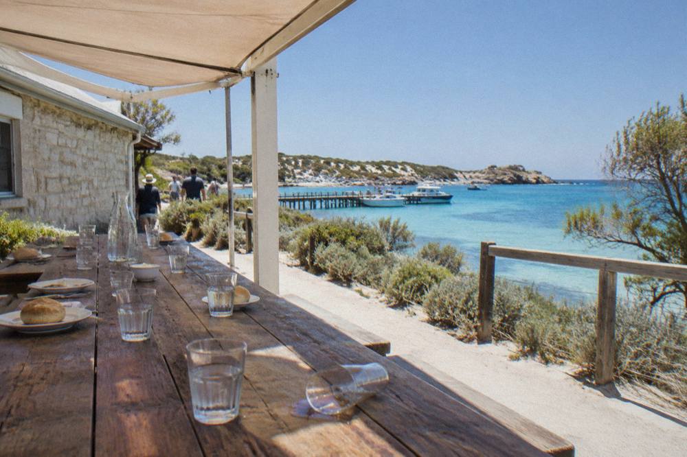 A long table set for lunch under a shaded veranda overlooking a blue bay