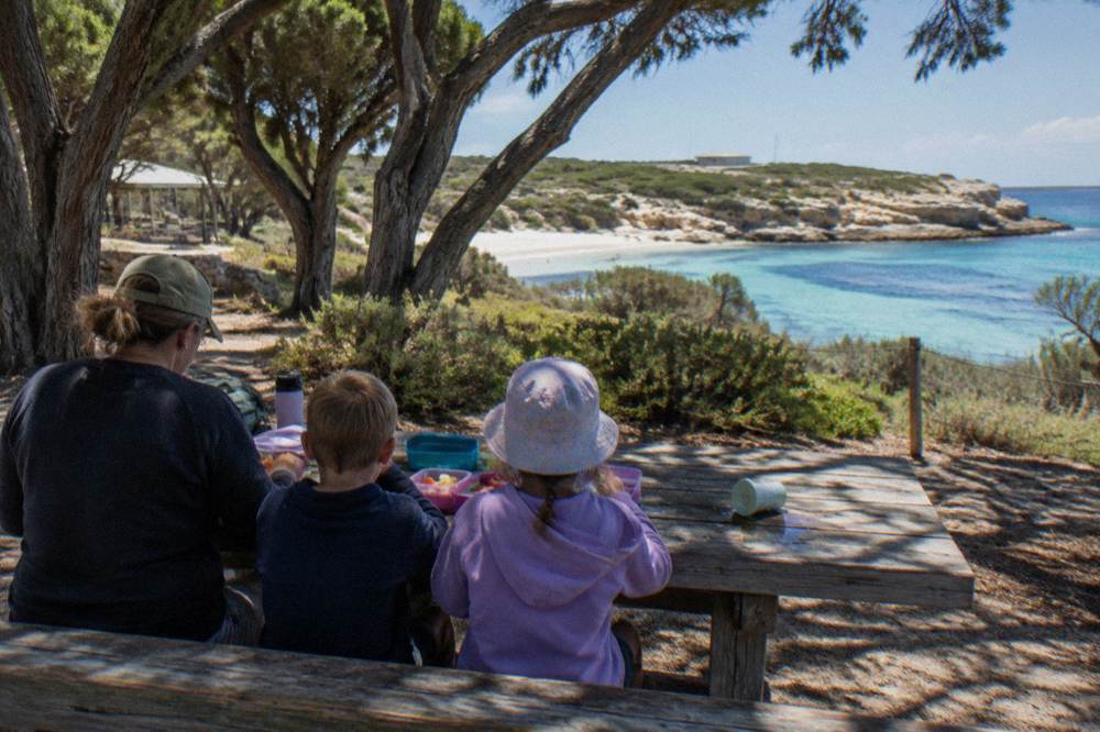 A family sharing food at a picnic table under trees near a coastal path