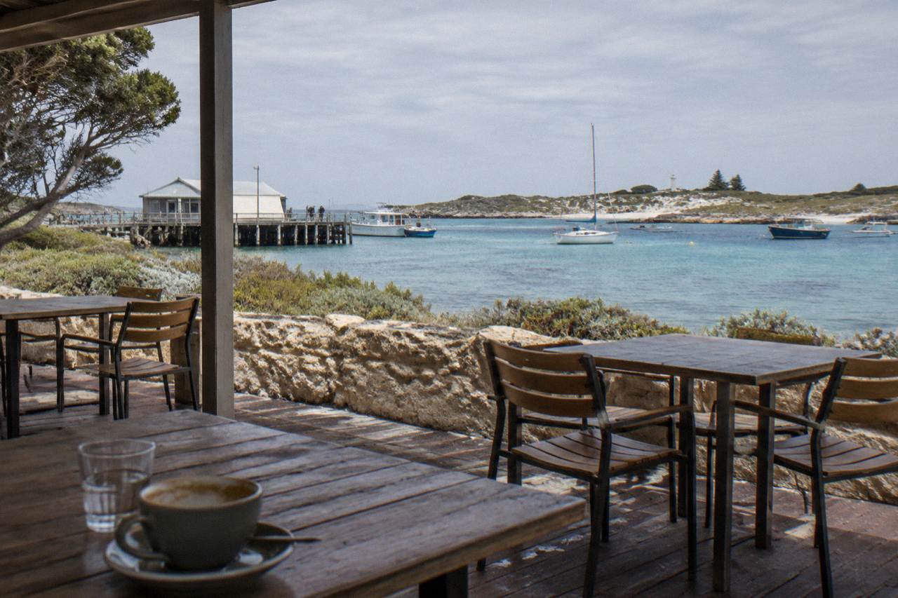 Outdoor tables beside a timber cafe veranda looking over a calm bay with moored boats