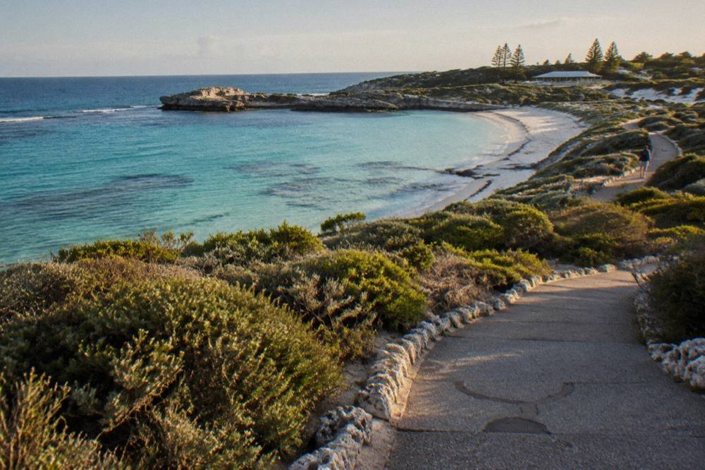 A paved shared path winding through low coastal scrub toward a turquoise bay
